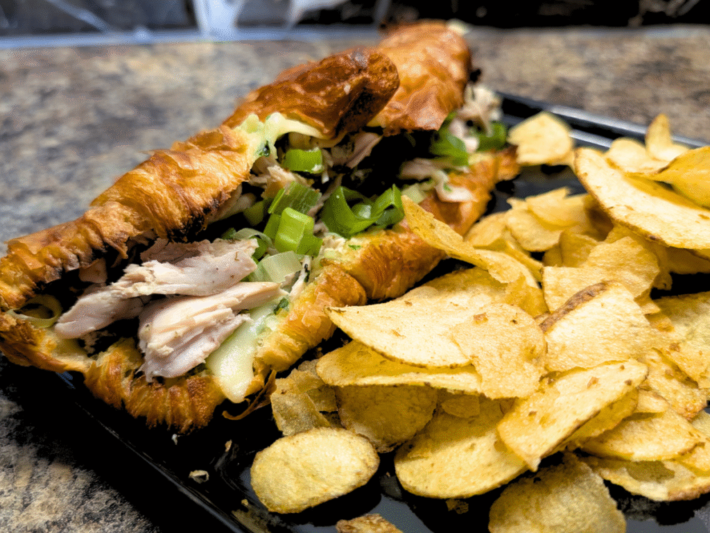 A croissant chicken grill made with leftover roast chicken served on a black plate with a side servings of crisps.